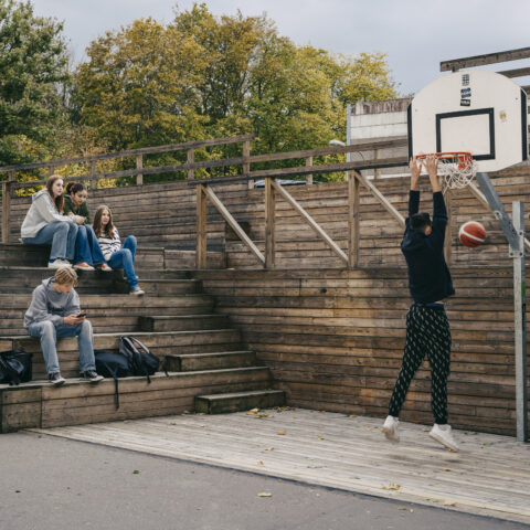 Skolgård med basketkorg, en tonårig kille dunkar en basketboll. På en läktare av trä sitter fyra personer.