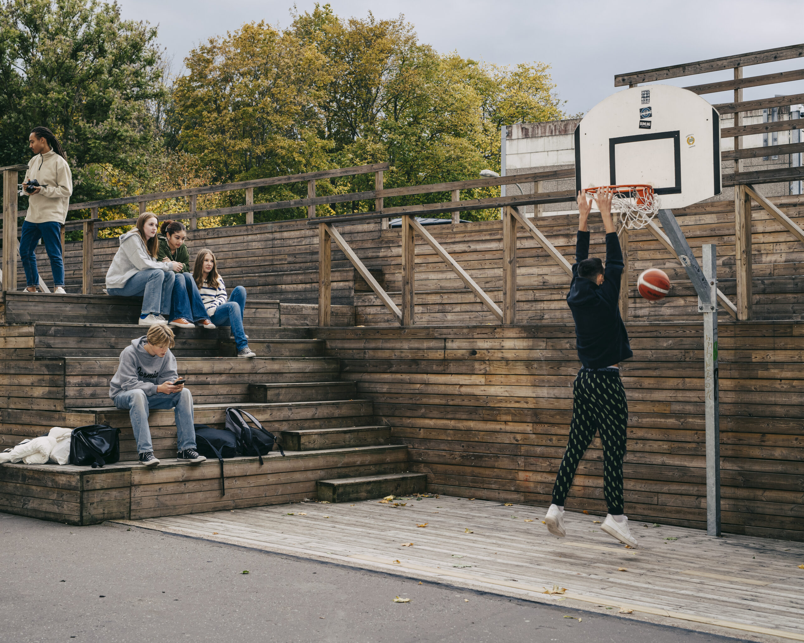 Skolgård med basketkorg, en tonårig kille dunkar en basketboll. På en läktare av trä sitter fyra personer.