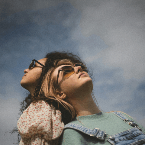 Två tjejer som står rygg mot rygg och en blå himmel med lätta slöjmoln