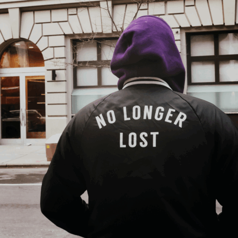 Ryggen på en person som står på gatan, luvan uppfälld och på jackan står det No longer lost