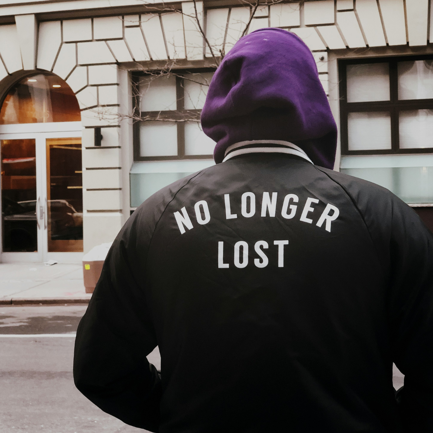 Ryggen på en person som står på gatan, luvan uppfälld och på jackan står det No longer lost