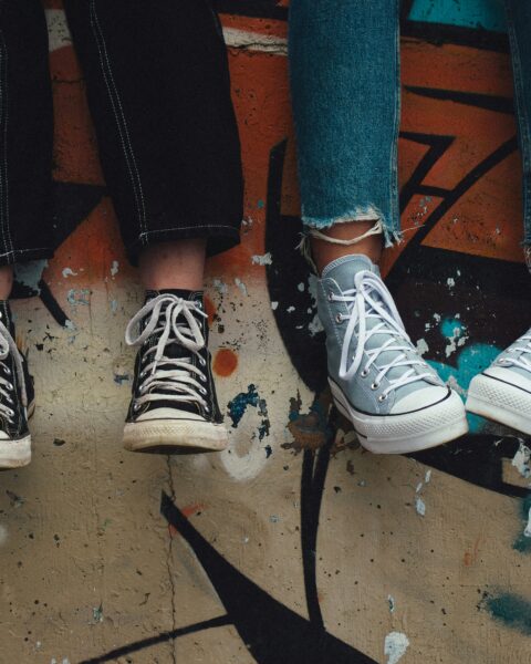 Två personer sitter på en graffiti-målad mur med dinglande ben. De bär slitna jeans och sneakers – ett par svarta och ett par ljusgråa Converse.